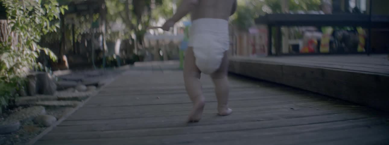 A baby is toddling across a porch in a backyard during the day