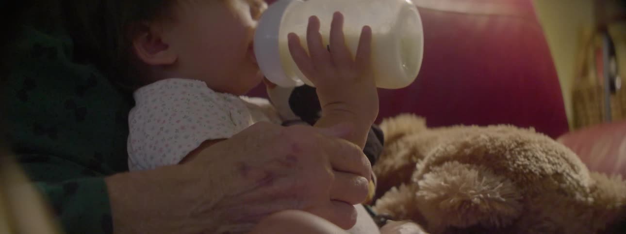 A baby drinks a bottle while sitting on an elderly woman's lap