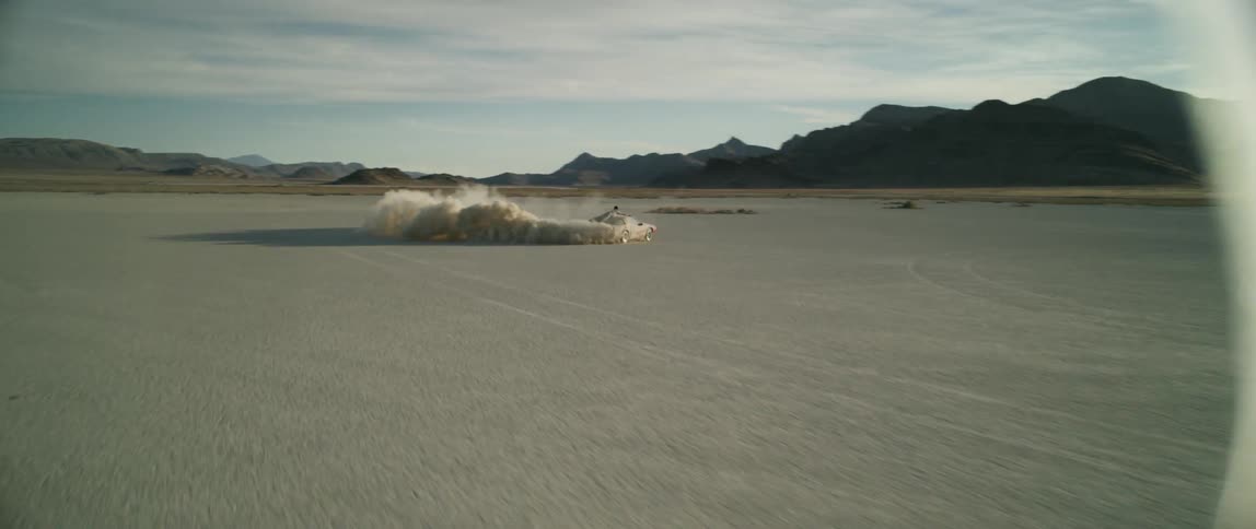 A man rids atop a roaring car as it does donuts on the desolate, dusty lakebed