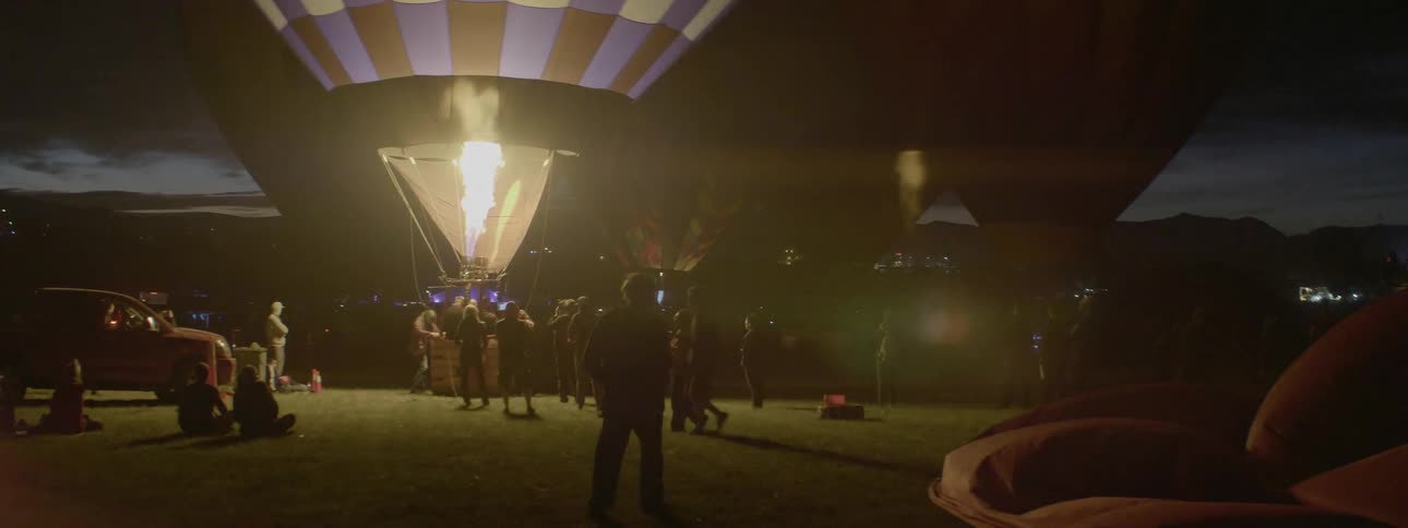 People mill about as hot air balloons are being prepared for flight at dusk