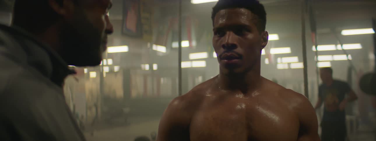 A trainer in a weight room talks to a man as he catches his breath