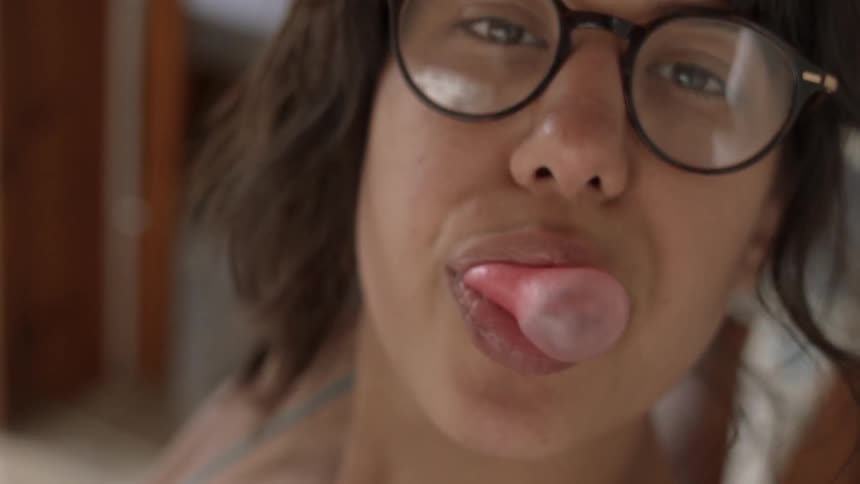 A woman in glasses is blowing bubbles with her chewing gum while looking at the camera