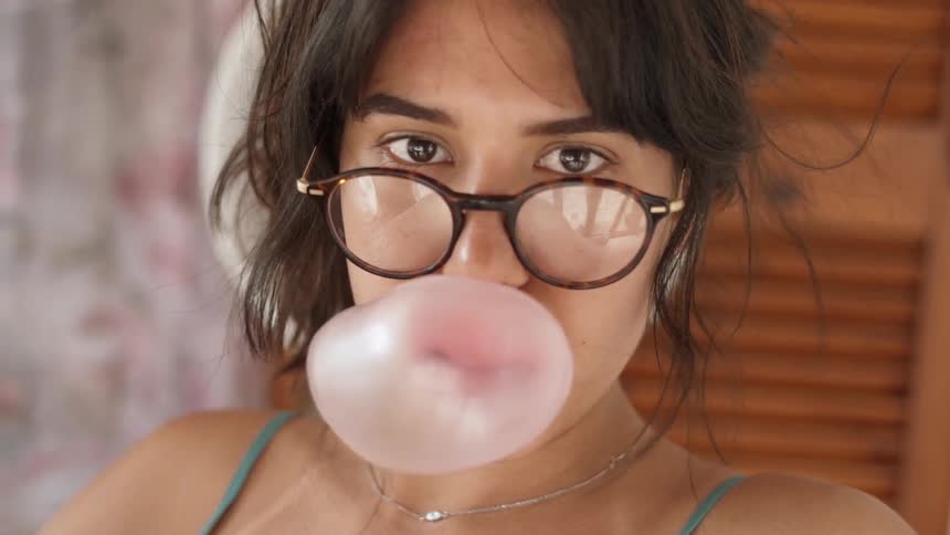 A woman wearing glasses blows a bubble with her gum
