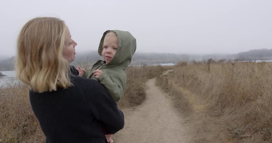 A woman holds a baby and walks down a dirt path by the water