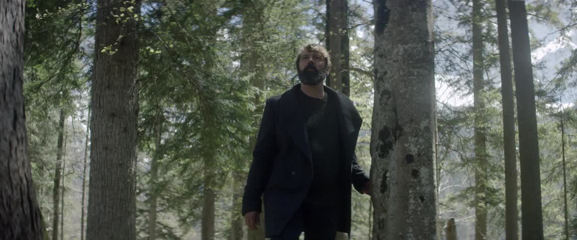 A man with a beard and jacket on is walking through the forest