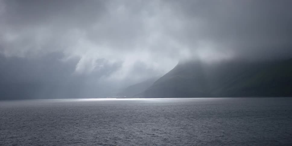 The sun is casting light rays through storm clouds as they pass over a coastline