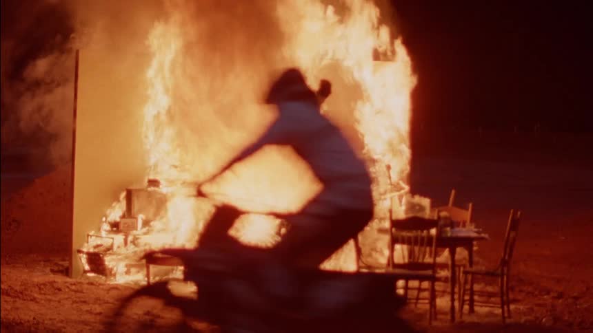 People ride dirt bikes around a pile of burning furniture