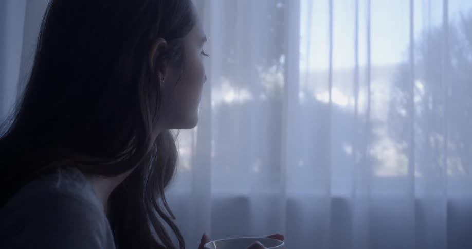 A woman holding a mug glances out the window