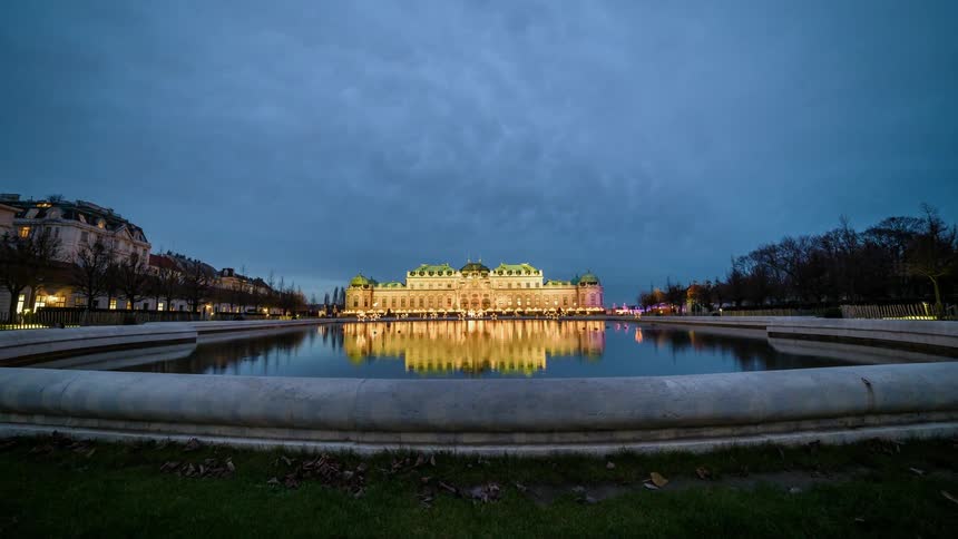 Clouds pass over a well lit mansion with a large pond in front