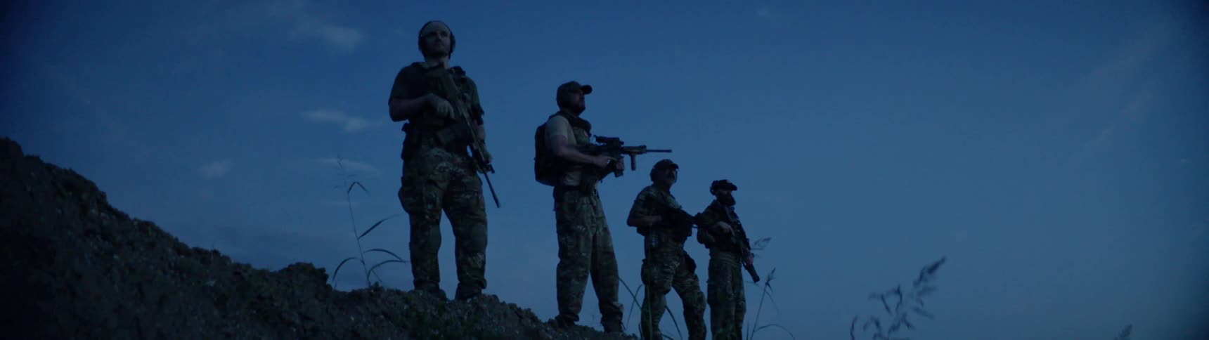 Four men in military garb and carrying guns are standing on a ridge at dusk