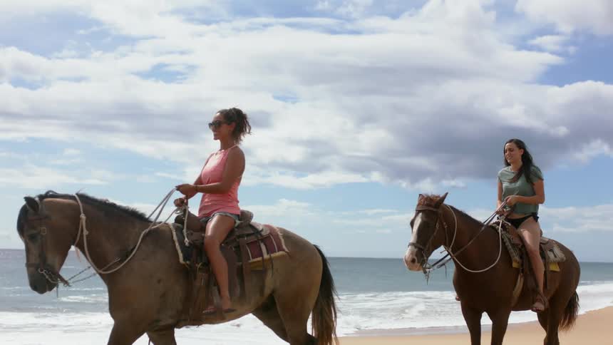 Two women smile and ride horses on the beach