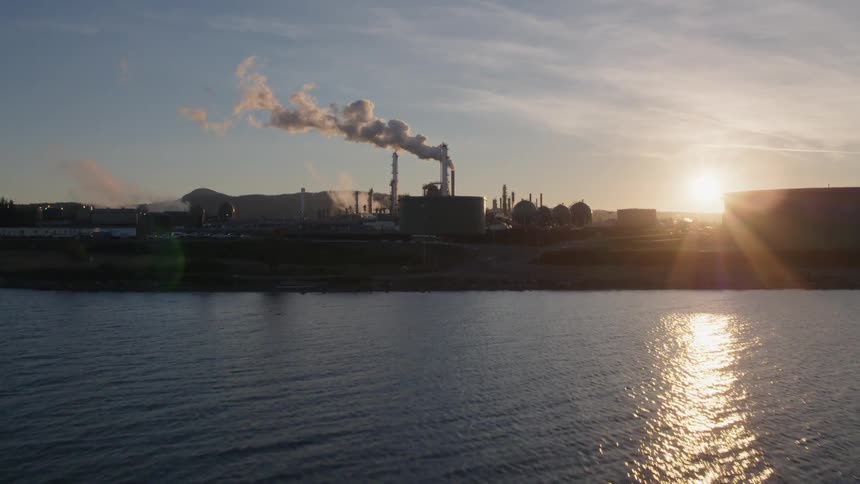 The sun peaks out from behind an oil tank while approaching a petroleum refinery near the ocean