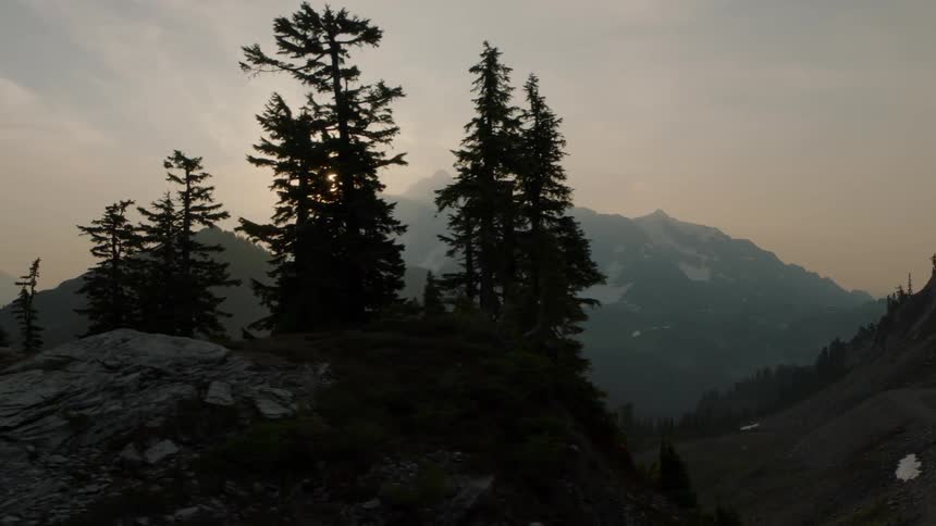 Aerial traverse behind a stand of silhouette fir trees as the sun rises from behind jagged mountain peaks