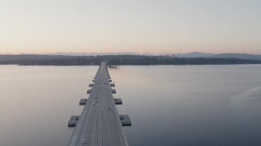 The sun rises over a long bridge crossing a calm lake with a city skyline and mountain range in the distance