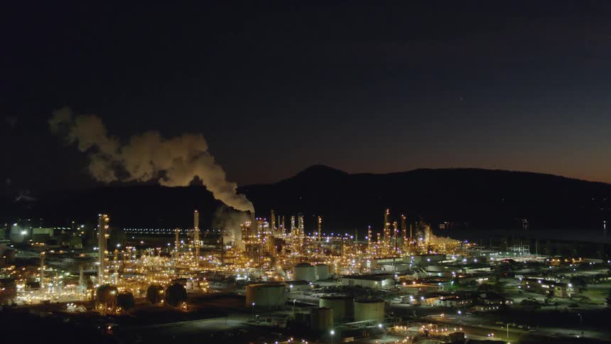 Rising above oil tanks as the refinery lights shimmer brightly at dusk