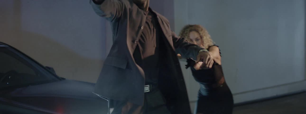 Man and woman jump off a car hood and the man aims a gun