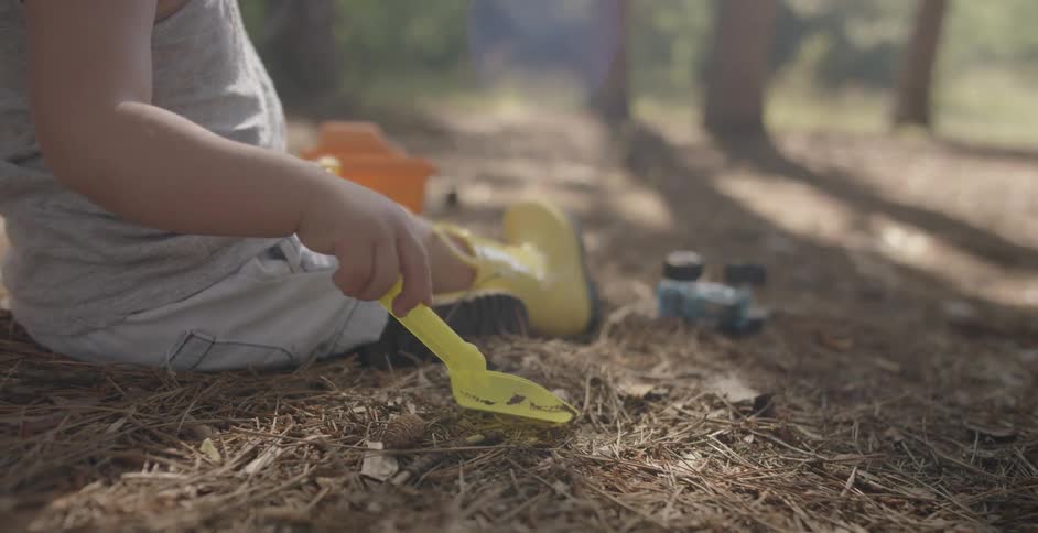 A toddler is using a toy shovel to dig in a forest floor on a sunny day