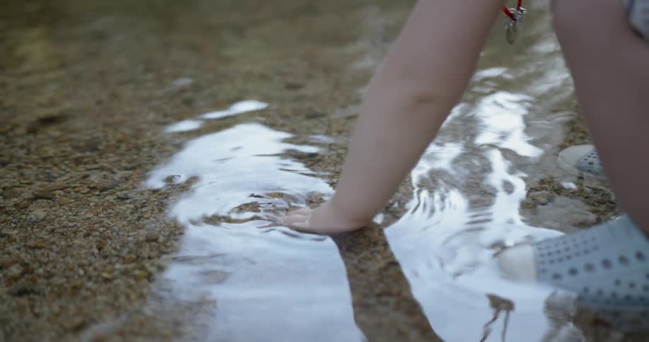 A toddler girl is kneeling down feeling water and sand in a stream