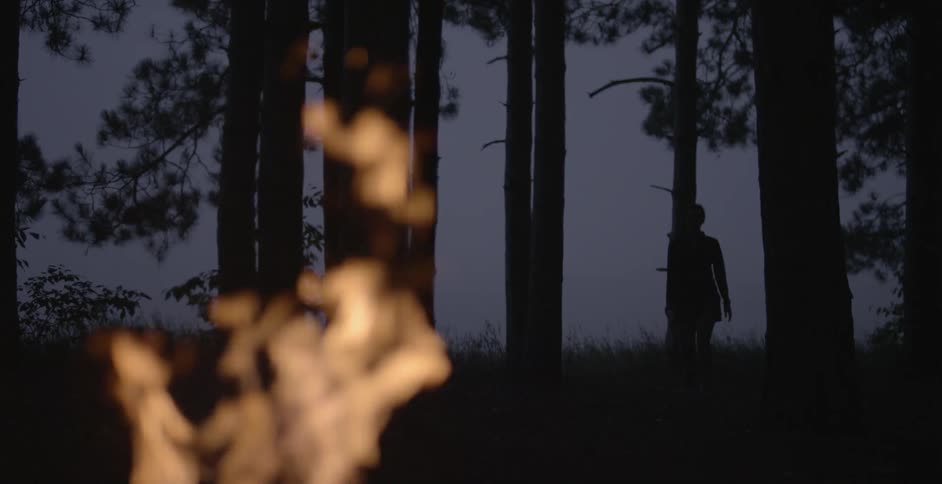 A silhouetted woman walks in a forest near a campfire
