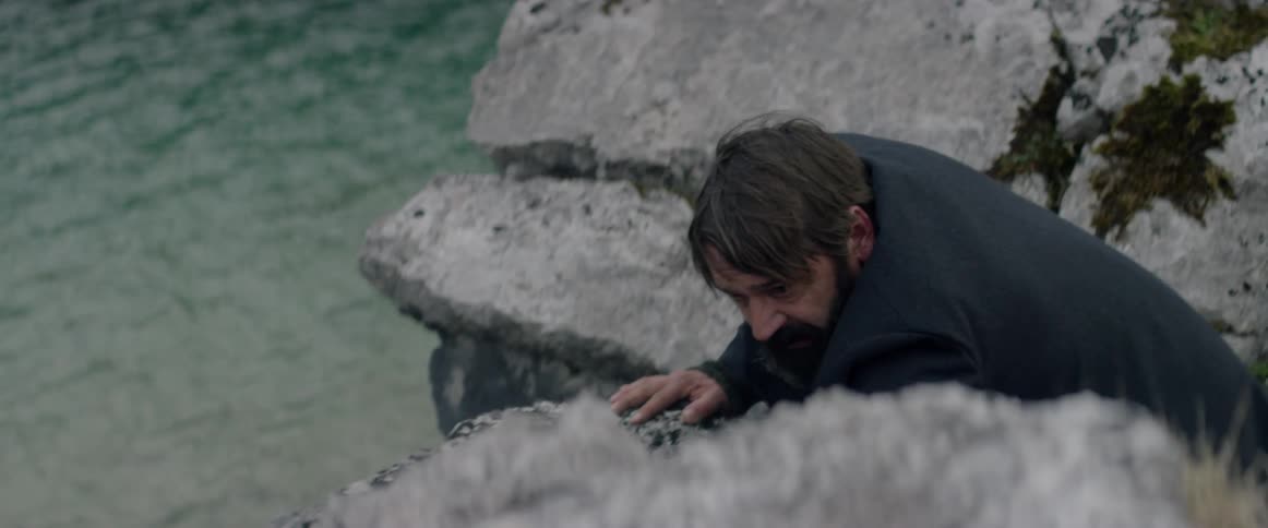 A man in an overcoat is crawling over boulders near the ocean during the day