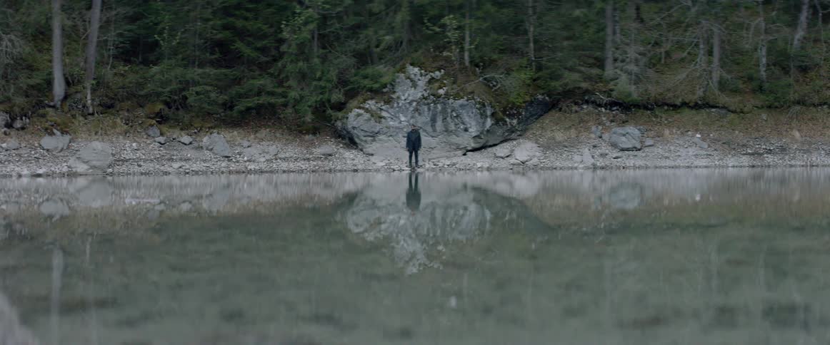 A man is standing at the edge of a lake looking at his reflection during the day