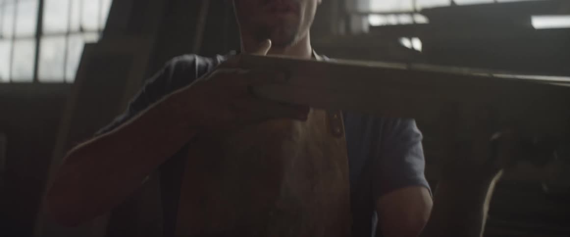 A man is looking at a piece of wood as he blows the dust off
