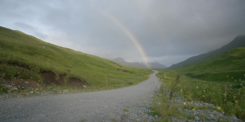 A rainbow is shining over a road in a natural setting on a stormy day