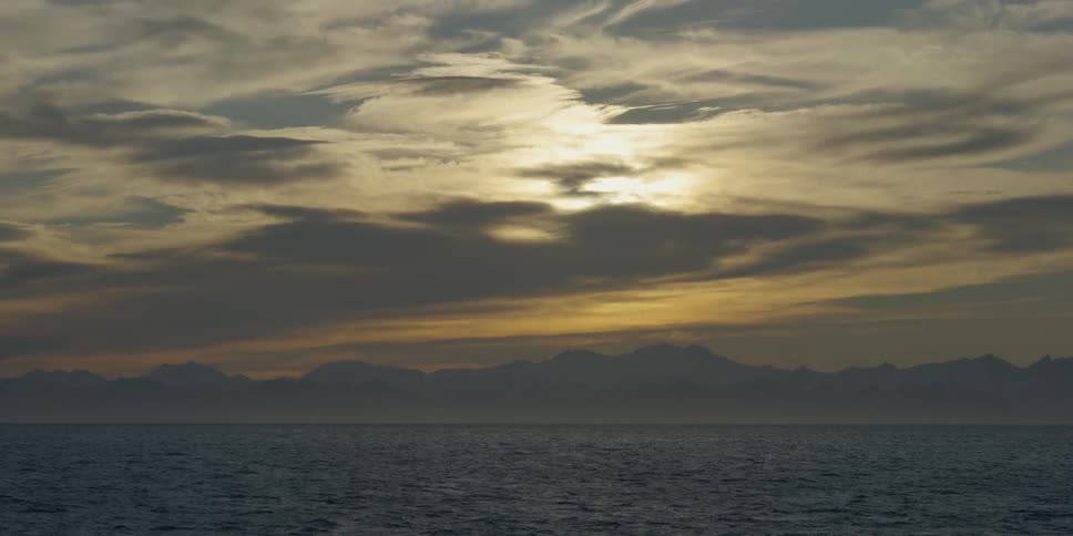 Clouds are covering the sun as it rises over coastal mountains