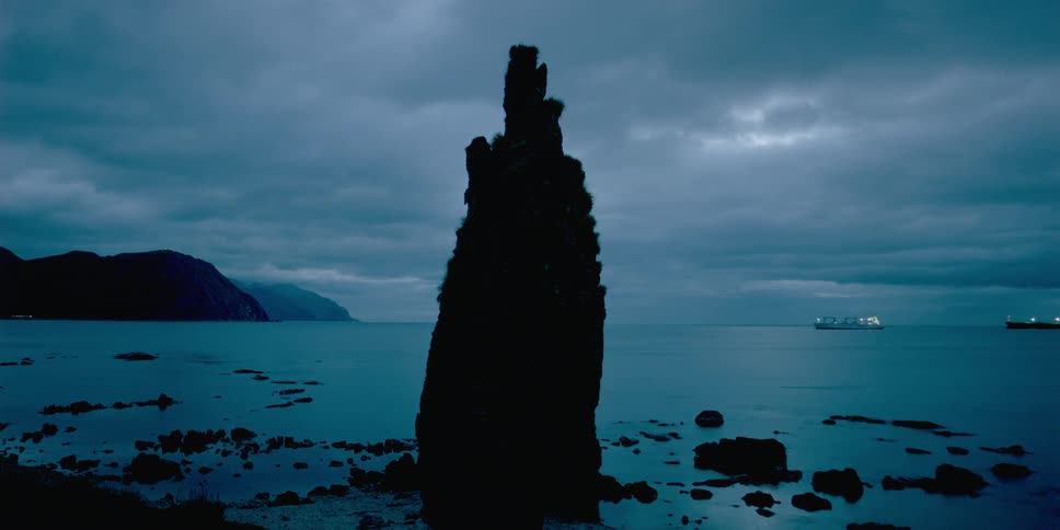 A rock stands tall in the water as a large ship floats