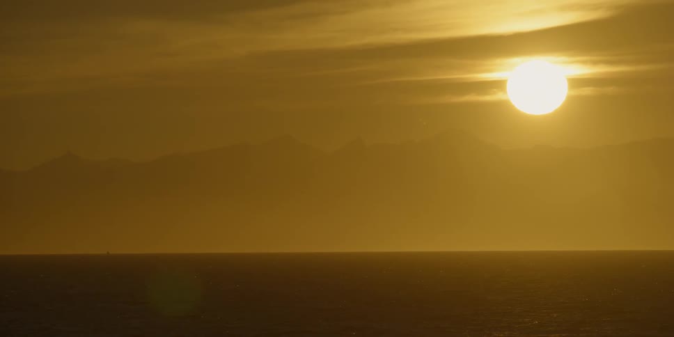 The sun is setting over the ocean near coastal mountains