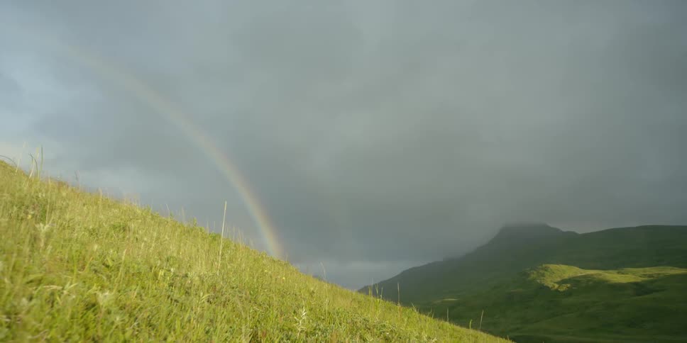A rainbow is shining beyond a hillside on a stormy day