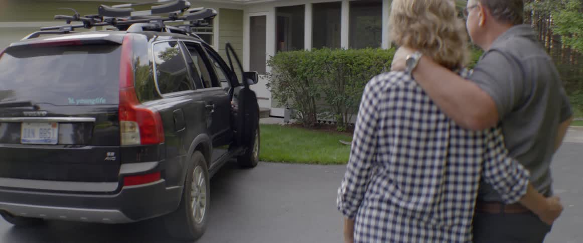 A couple wave goodbye as a young man gets in a car