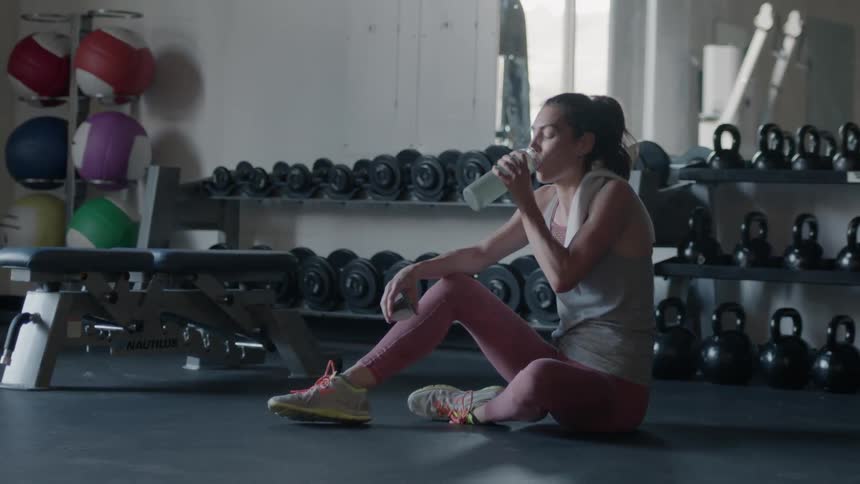 A woman grabs a water bottle and rests in a gym