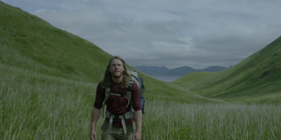 Man wearing a large backpack walks through tall grass near hills