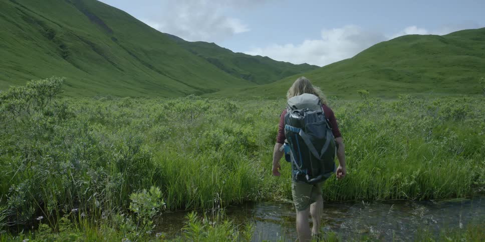 A hiker crosses a stream in a grassy valley