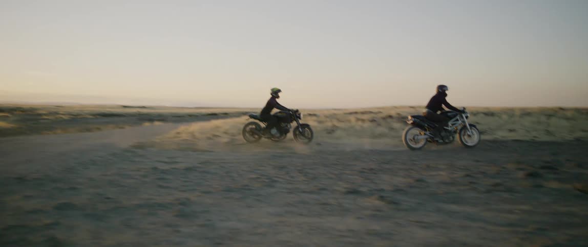 Two people ride motorbikes on a dirt path at sunset