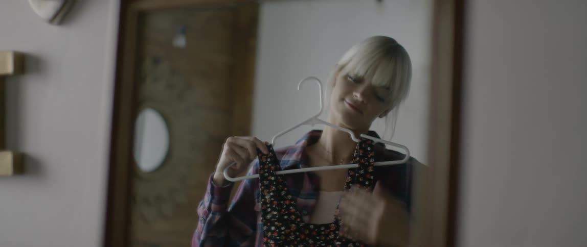 A blond haired woman holds a dress up to her and looks in the mirror