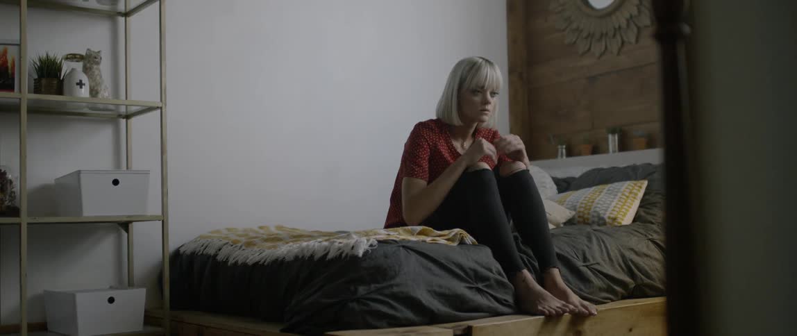Woman sits at the edge of a bed curled up and frowning