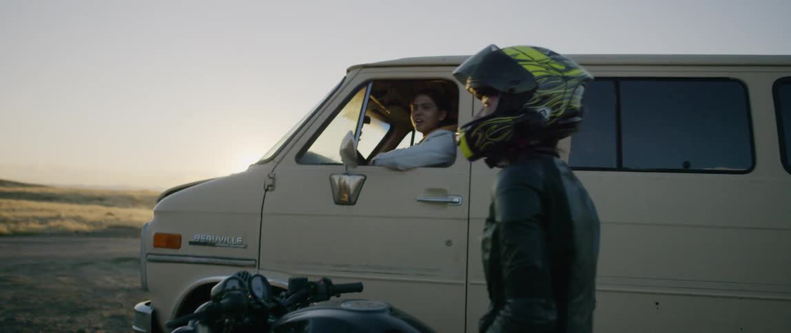 A van pulls up next to a motorcyclist in a rural field at sunset