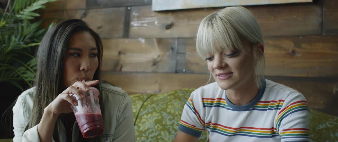 Two women sit on a couch and drink smoothies
