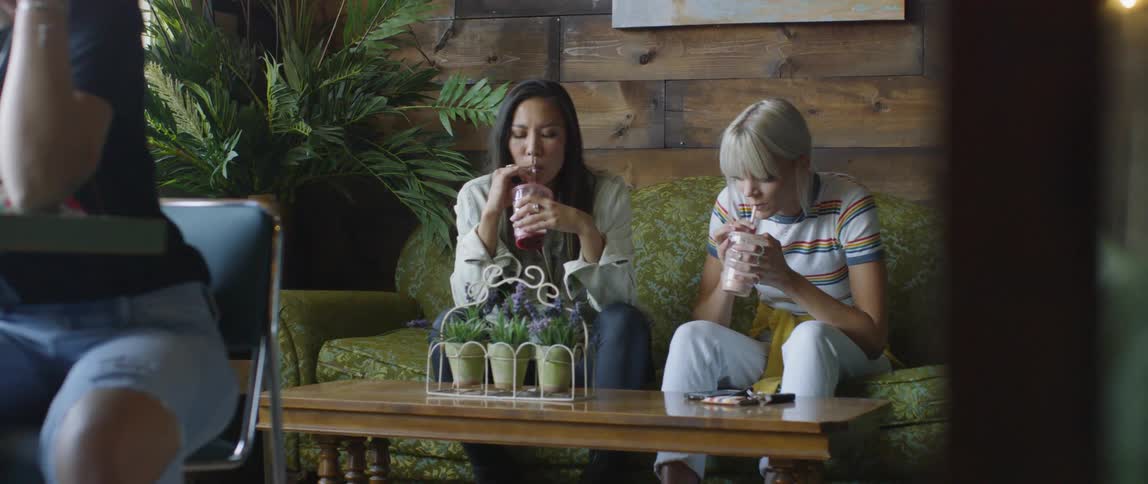 Two women sip smoothies while sitting on a couch