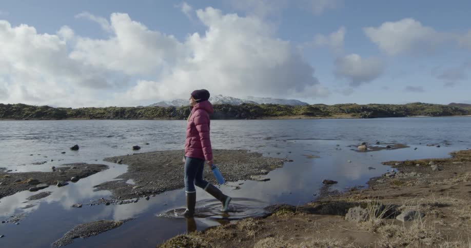 A woman in a pink jacket and rain boots walks across a river
