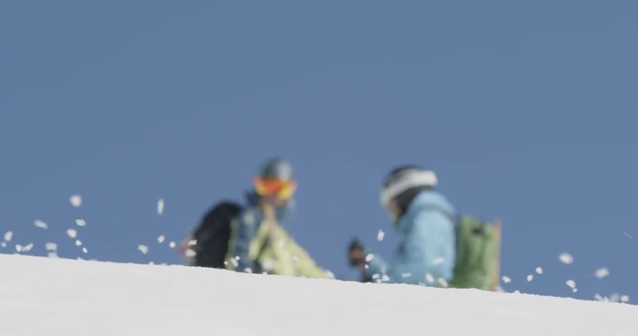 Snow is drifting downhill in front of two skiers on a sunny day