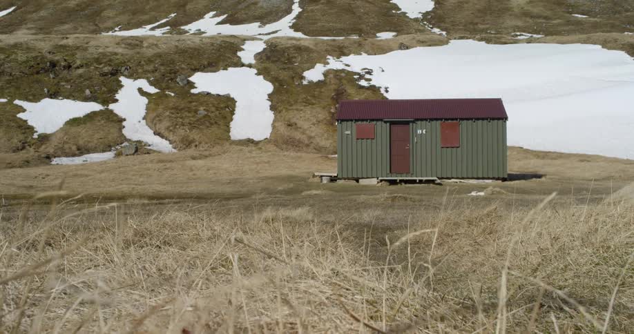 The wind is blowing the grass outside a cabin in a natural setting during the day