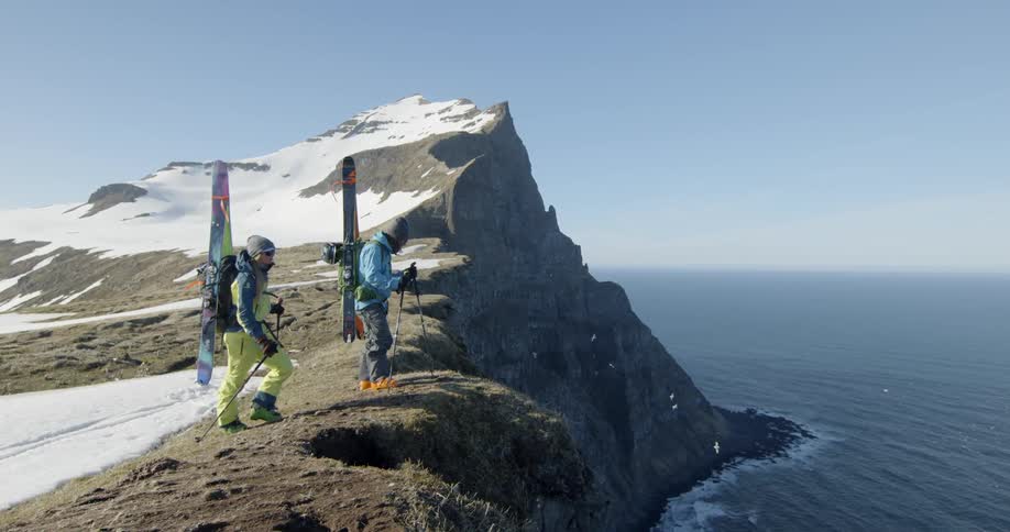 Two skiers stop and look over the edge of a cliff to see the ocean below