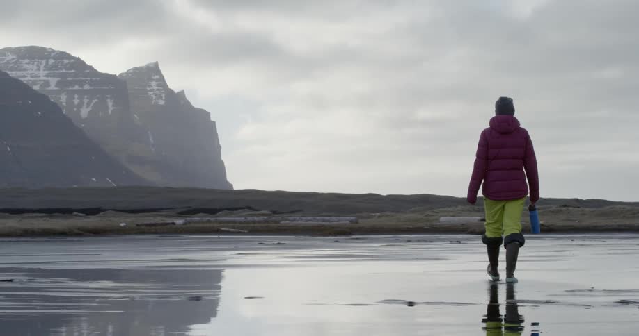 A woman is carrying a water bottle as she walks on a beach on a cold day