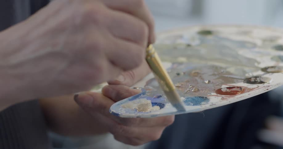 An artist dips his brush in paint on a palette