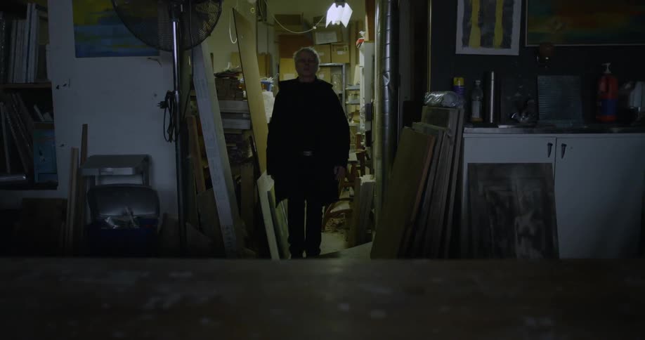 Man in a trench coat turns light on and walks through cluttered basement