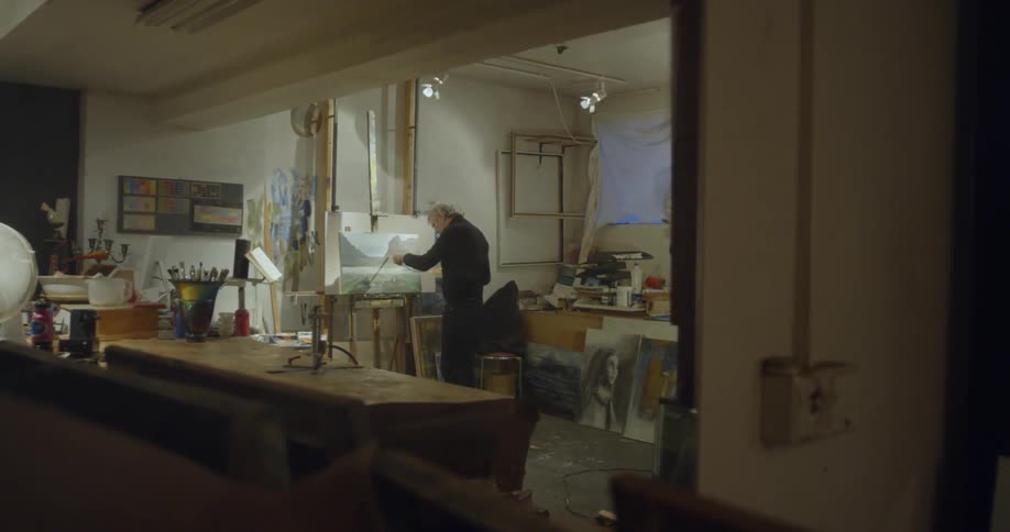 An artist paints on a canvas in cluttered art studio
