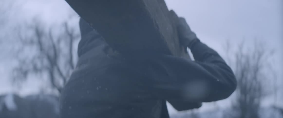 A man is picking up a large log in the snow and puts it over his shoulder